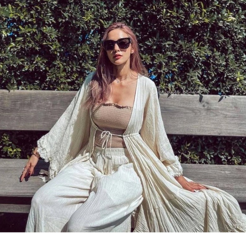 Op de afbeelding: Een vrouw draagt een beige tube top, een bijpassende broek en een lange, cr&egrave;mekleurige open cardigan. Ze draagt ook een zwarte zonnebril en zit op een houten bank voor een groene, bladerrijke achtergrond.