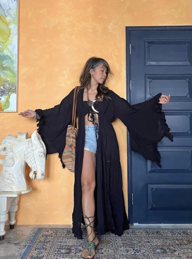 May include: A long, flowing black robe with wide sleeves and a tie closure. The robe is worn over a black top and denim shorts. A patterned tote bag hangs from the shoulder. The outfit is accessorized with sandals.