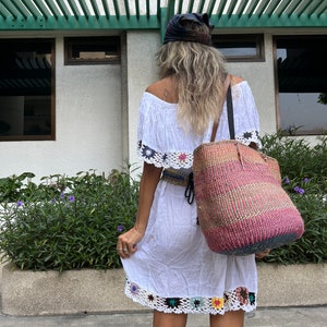 May include: A woman wearing a white off-the-shoulder dress with crochet trim and a black headband. She is carrying a large, colorful woven tote bag.