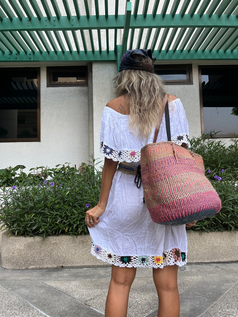 May include: A woman wearing a white off-the-shoulder dress with crochet trim and a black headband. She is carrying a large, colorful woven tote bag.