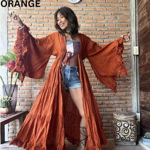 May include: An orange, long-sleeved robe with ruffled sleeves and a tie waist. The word "ORANGE" is displayed in white block letters. The robe is worn over a patterned top and denim shorts. The background is a brick wall.