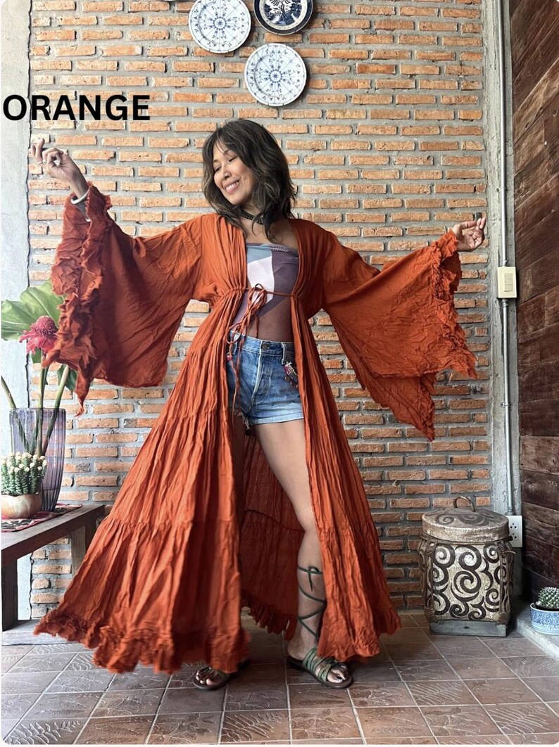 May include: An orange, long-sleeved robe with ruffled sleeves and a tie waist. The word "ORANGE" is displayed in white block letters. The robe is worn over a patterned top and denim shorts. The background is a brick wall.