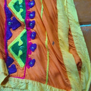 May include: A close-up of a bright orange fabric with colorful embroidery. The fabric has a yellow border and is decorated with green, purple, and pink stitching.