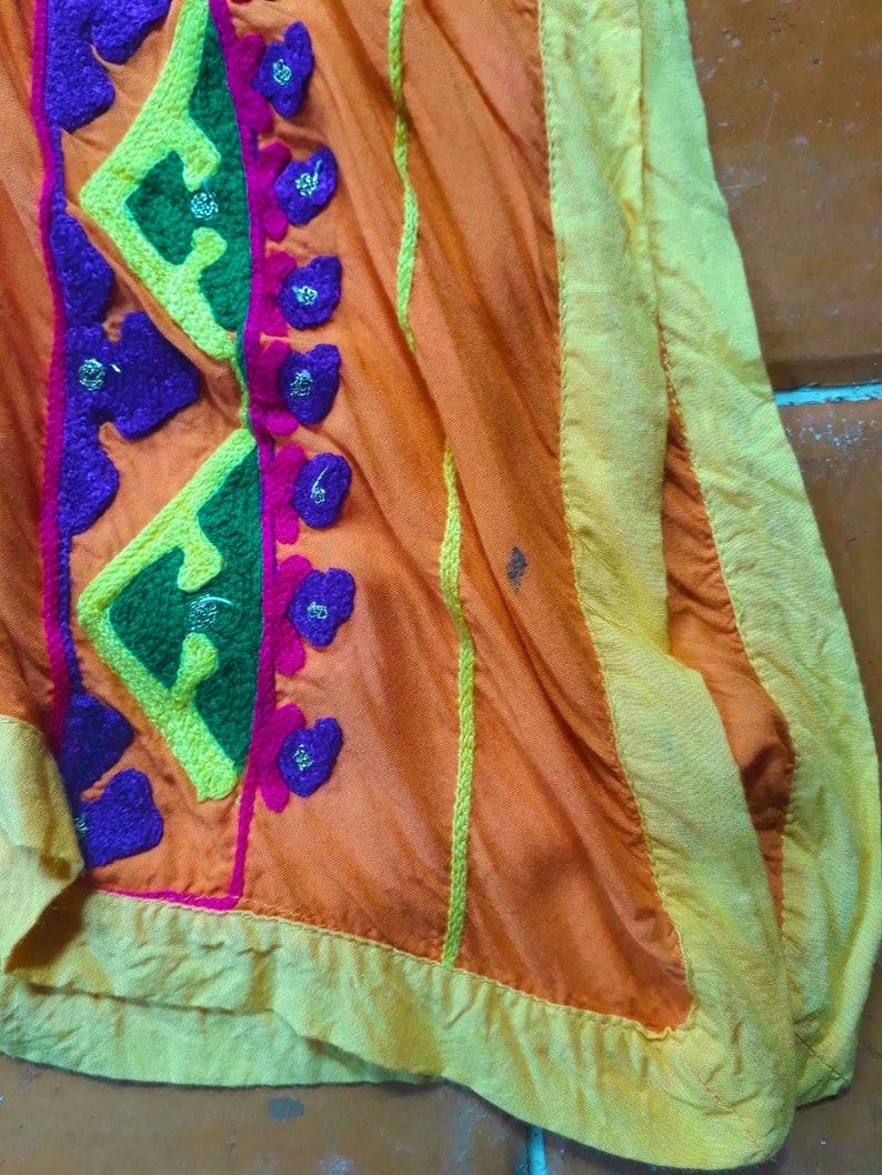 May include: A close-up of a bright orange fabric with colorful embroidery. The fabric has a yellow border and is decorated with green, purple, and pink stitching.