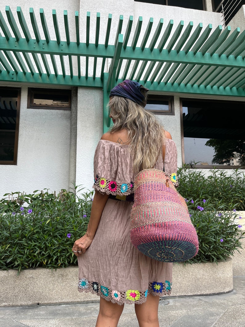 May include: A woman wearing a brown crochet dress with colorful floral accents and a large, woven, multi-colored tote bag.