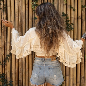May include: A woman wearing a white crochet top with a ruffled hem and a pair of light blue denim shorts with frayed edges. She is standing in front of a bamboo wall.