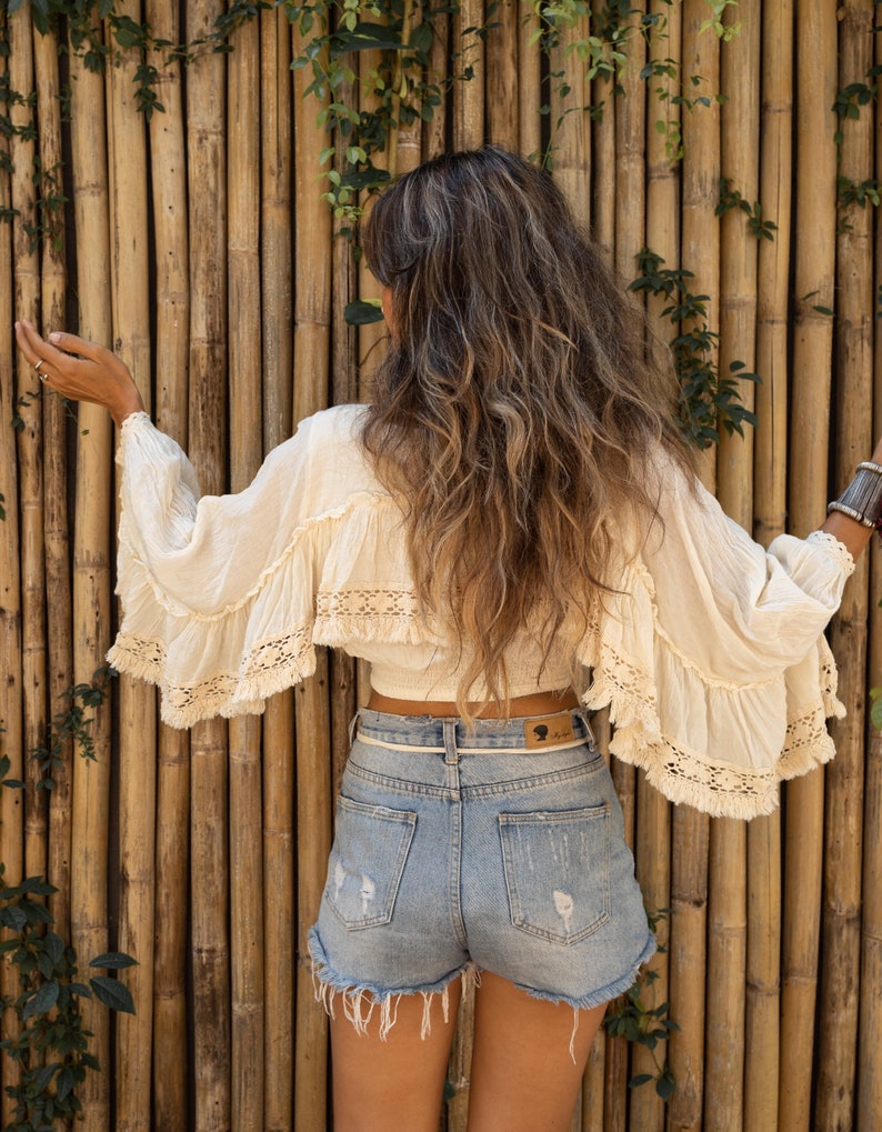 May include: A woman wearing a white crochet top with a ruffled hem and a pair of light blue denim shorts with frayed edges. She is standing in front of a bamboo wall.