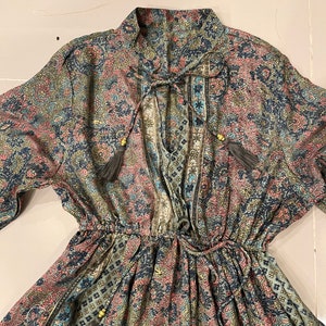May include: A long-sleeved dress with a paisley print in shades of blue, green, pink, and gold. The dress has a V-neckline, a tie at the neckline, and a drawstring waist.