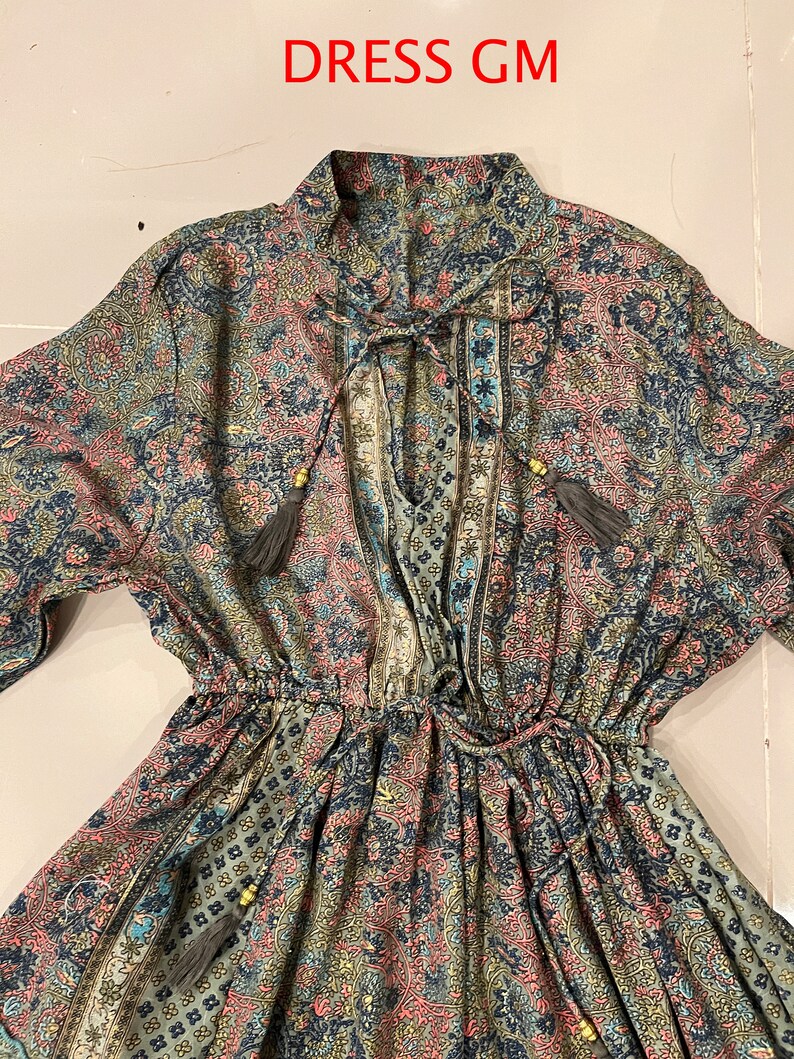 May include: A long-sleeved dress with a paisley print in shades of blue, green, pink, and gold. The dress has a V-neckline, a tie at the neckline, and a drawstring waist.