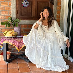 May include: A woman wearing a white, flowing, tiered dress with lace detailing. She is sitting on a wooden floor with a table in front of her. The table has a colorful tablecloth and a bowl of fruit on it.