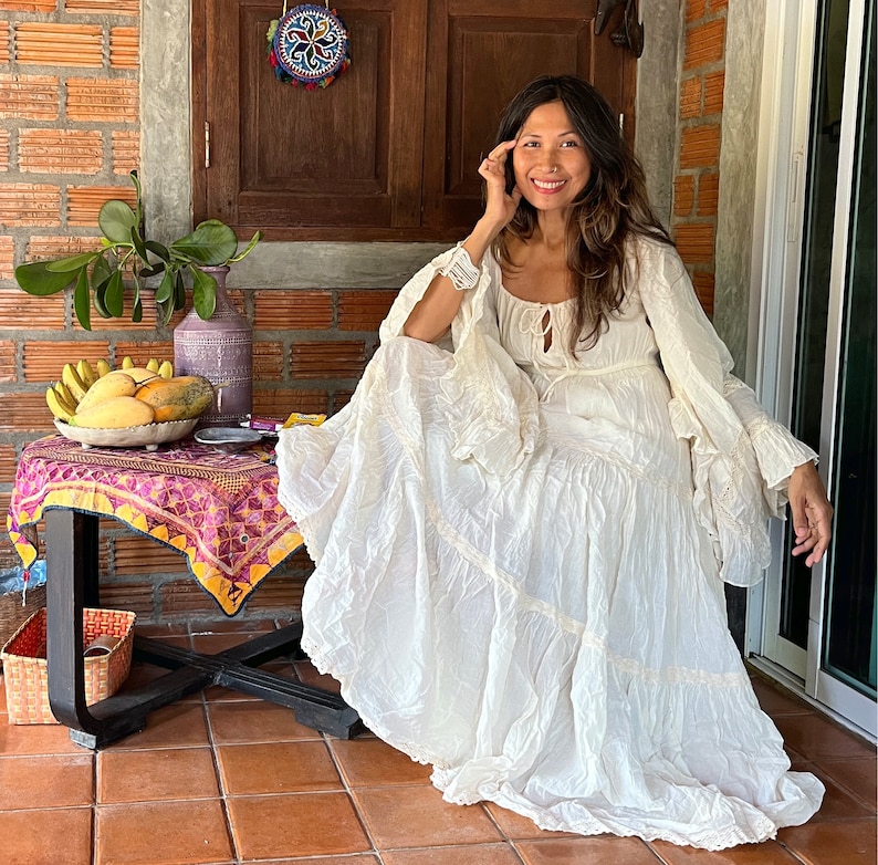 May include: A woman wearing a white, flowing, tiered dress with lace detailing. She is sitting on a wooden floor with a table in front of her. The table has a colorful tablecloth and a bowl of fruit on it.