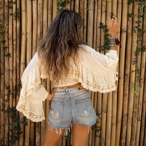 May include: A woman wearing a white crochet top with a brown belt that says "Spellmaya" and denim shorts. She is standing in front of a bamboo wall.