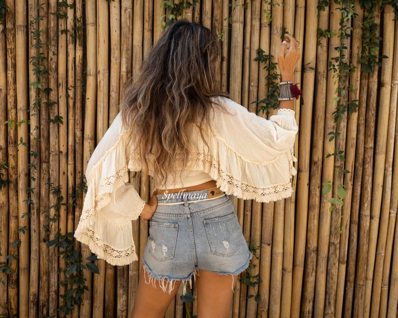 May include: A woman wearing a white crochet top with a brown belt that says "Spellmaya" and denim shorts. She is standing in front of a bamboo wall.