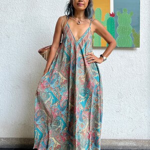 May include: A woman wearing a long, flowing, blue and brown paisley print maxi dress with a V-neckline. She is standing in front of a white wall with a painting of cacti behind her.