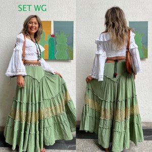 May include: A green tiered skirt with lace trim and a white peasant top with a lace trim. The skirt has a brown leather belt with a silver buckle. The top has a brown leather strap with a silver buckle. The model is standing in front of a wall with a painting of cacti on it. The text "SET WG" is written in green letters at the top of the image.