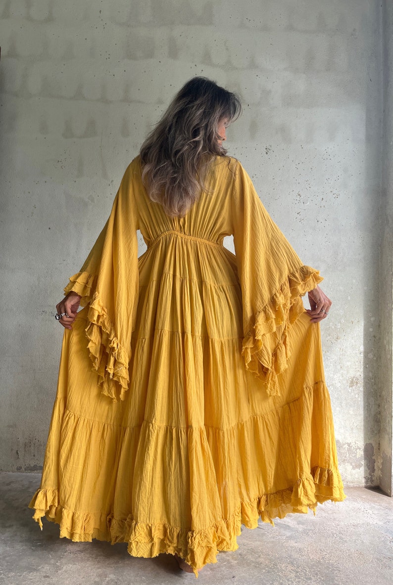 May include: A long, flowing mustard yellow dress with tiered skirt and ruffled bell sleeves. The dress has a cinched waist and a full, floor-length skirt. The fabric appears to be lightweight and textured, suitable for warm weather.