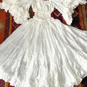 May include: White off-the-shoulder peasant top with ruffled sleeves and a white tiered skirt. The set is made of a lightweight fabric and has a flowy, romantic style. The text "Set White" is visible in the image.
