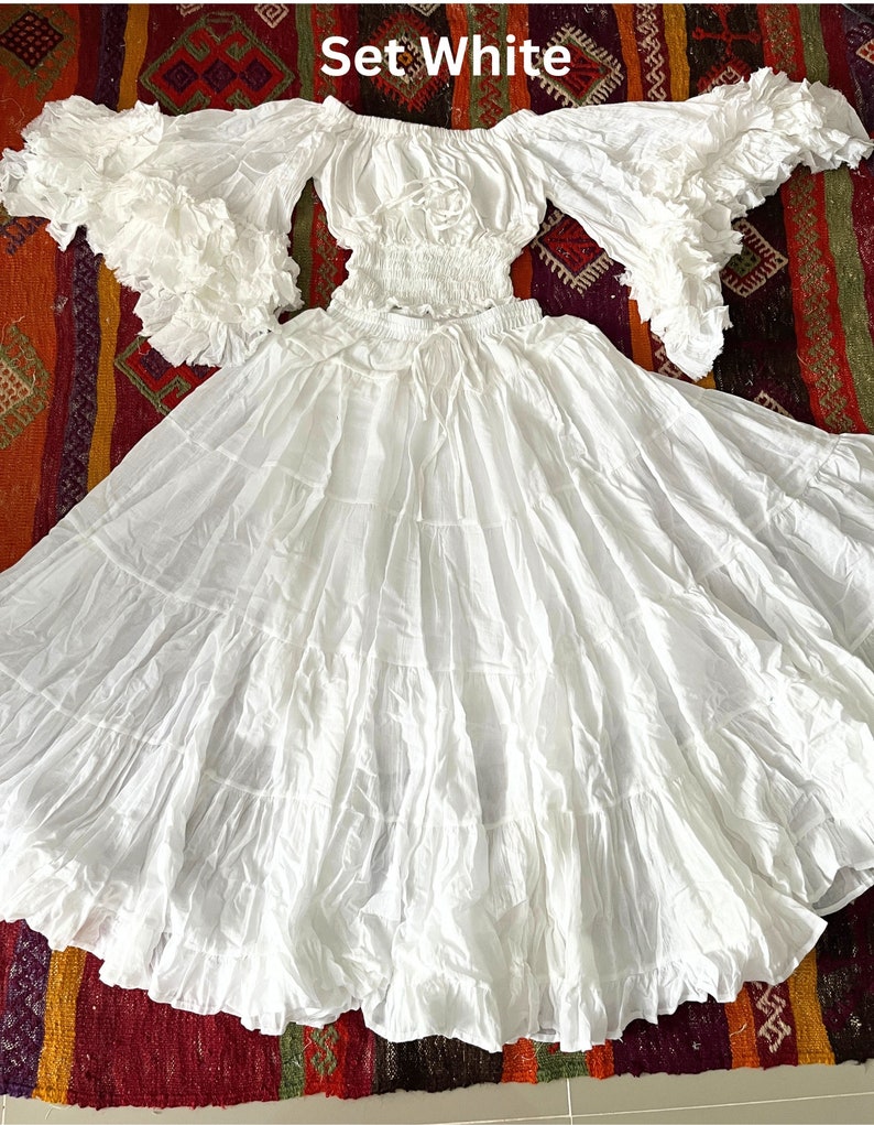 May include: White off-the-shoulder peasant top with ruffled sleeves and a white tiered skirt. The set is made of a lightweight fabric and has a flowy, romantic style. The text "Set White" is visible in the image.
