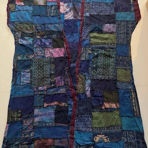 May include: A sleeveless open-front vest featuring a patchwork design composed of various blue, purple, and green fabric squares with diverse patterns. The garment includes a dark maroon trim along the front edges and displays the text Style BU3.