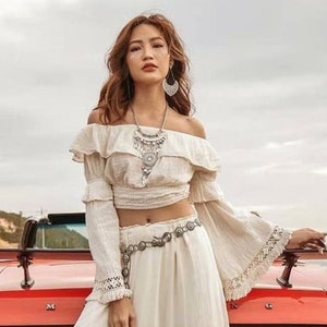 May include: A woman wearing a white off-the-shoulder top with a ruffled neckline and long sleeves. She is wearing a silver necklace and a silver belt. The top is cropped and shows her midriff. She is standing in front of a red car.