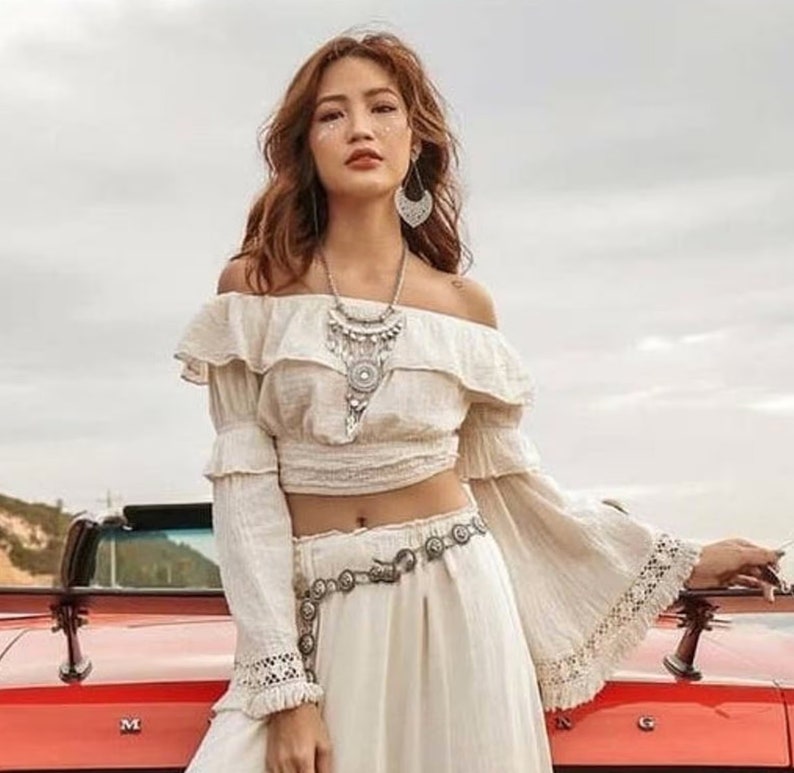 May include: A woman wearing a white off-the-shoulder top with a ruffled neckline and long sleeves. She is wearing a silver necklace and a silver belt. The top is cropped and shows her midriff. She is standing in front of a red car.