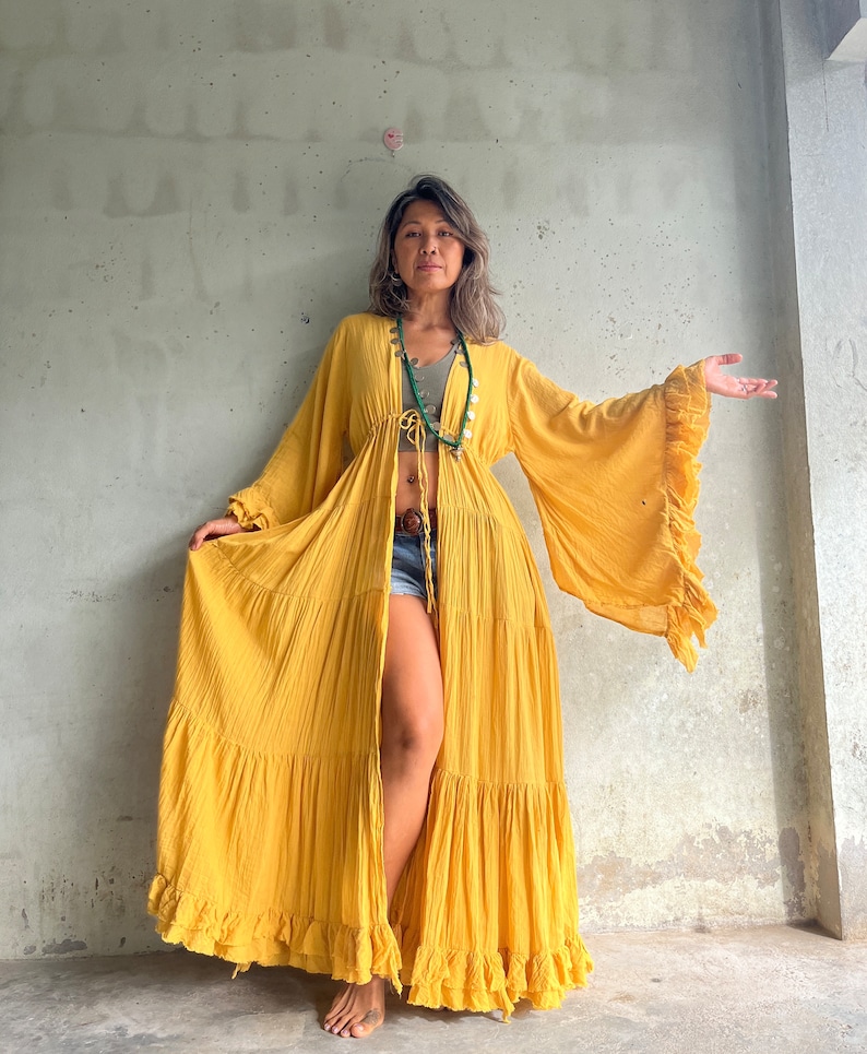 May include: A long, flowing mustard yellow robe with bell sleeves and a ruffled hem. The robe is open in the front and tied at the waist. The sleeves have a ruffled edge. The robe is made of a lightweight fabric.