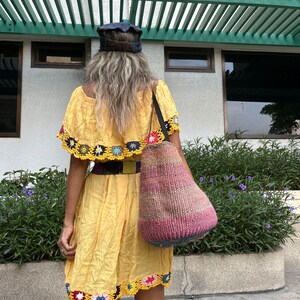 May include: A woman wearing a yellow crochet-trimmed dress and a blue cap carries a woven tote bag with colorful stripes.