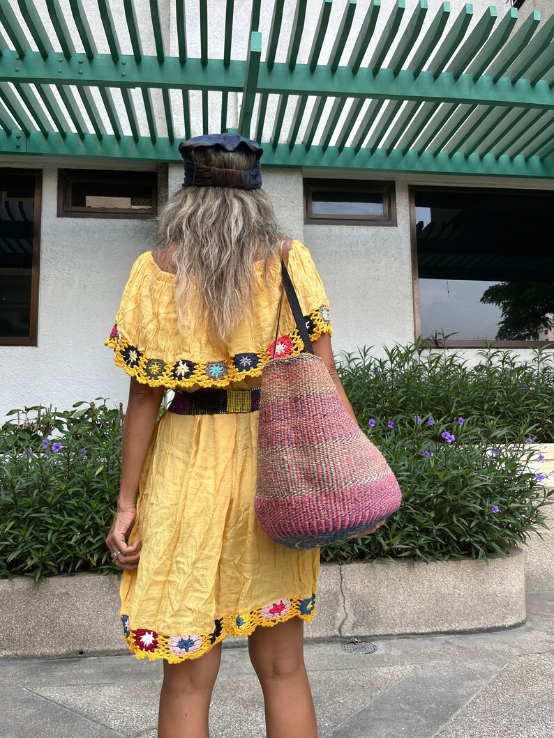 May include: A woman wearing a yellow crochet-trimmed dress and a blue cap carries a woven tote bag with colorful stripes.