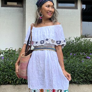 May include: A white off-the-shoulder dress with crochet trim and a colorful beaded belt. The dress has a ruffled neckline and a flowy skirt. The model is wearing a blue bandana and large hoop earrings.