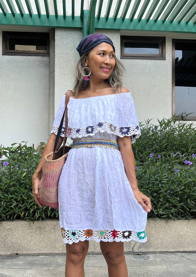 May include: A white off-the-shoulder dress with crochet trim and a colorful beaded belt. The dress has a ruffled neckline and a flowy skirt. The model is wearing a blue bandana and large hoop earrings.