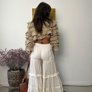Pode incluir: Um top cropped bege com babados e cal&ccedil;as palazzo brancas em camadas. O top tem v&aacute;rias camadas de babados, e as cal&ccedil;as t&ecirc;m um design de perna larga com acabamento em renda. O conjunto faz parte de uma cole&ccedil;&atilde;o de roupas de estilo bo&ecirc;mio.