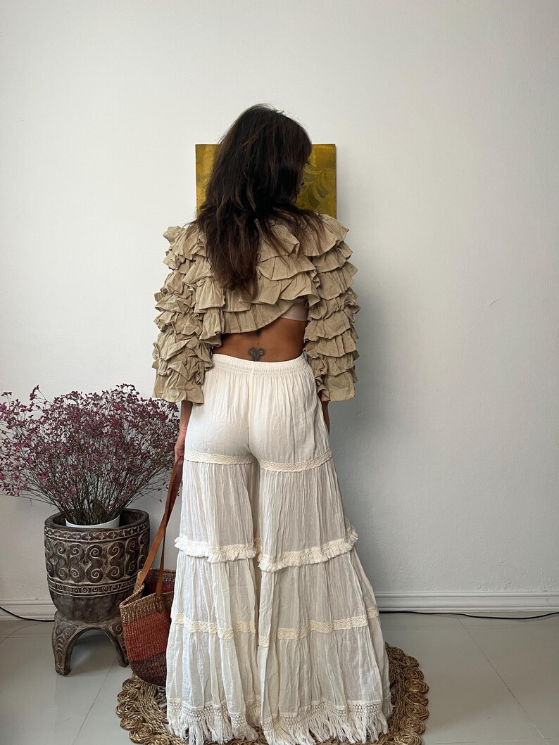 Pode incluir: Um top cropped bege com babados e cal&ccedil;as palazzo brancas em camadas. O top tem v&aacute;rias camadas de babados, e as cal&ccedil;as t&ecirc;m um design de perna larga com acabamento em renda. O conjunto faz parte de uma cole&ccedil;&atilde;o de roupas de estilo bo&ecirc;mio.