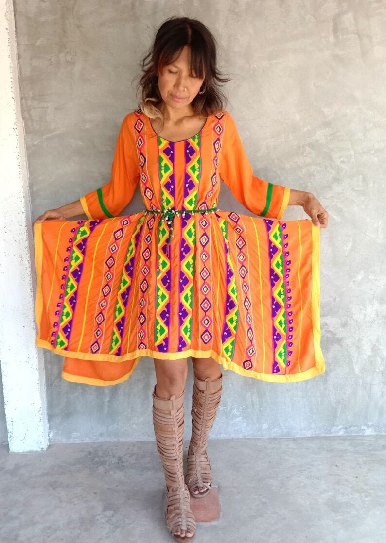 May include: A bright orange dress with a colorful geometric pattern. The dress has a high neckline and long sleeves. The dress is made of a lightweight fabric and has a flowing silhouette.