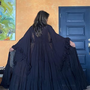 Op de afbeelding: Een lange, vloeiende zwarte jurk met gelaagde ruches en wijde mouwen. De jurk heeft een doorschijnend paneel op de rug en is gemaakt van een lichte stof. Het model staat voor een oranje muur en een blauwe deur.