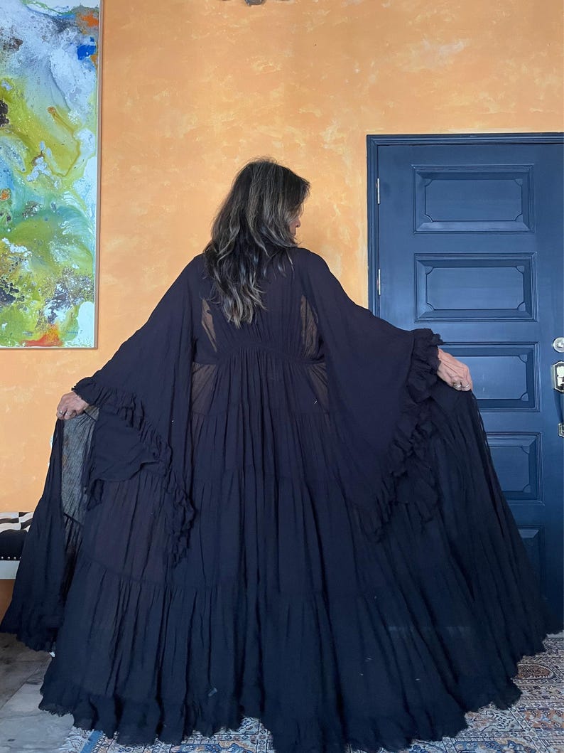 Op de afbeelding: Een lange, vloeiende zwarte jurk met gelaagde ruches en wijde mouwen. De jurk heeft een doorschijnend paneel op de rug en is gemaakt van een lichte stof. Het model staat voor een oranje muur en een blauwe deur.