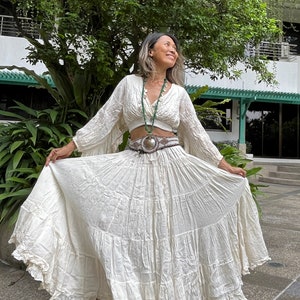 May include: A white, flowing maxi skirt with multiple tiers and a ruffled hem. The skirt is paired with a white, cropped, long-sleeved blouse. The model is wearing a silver belt with a decorative buckle.