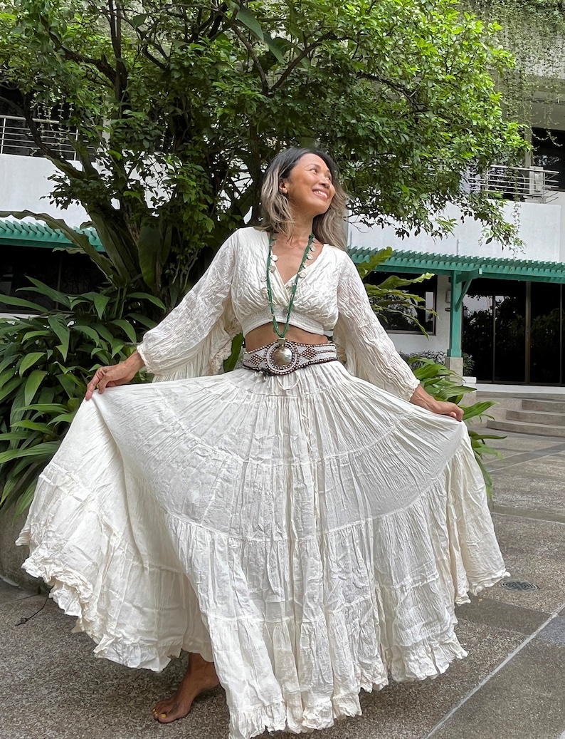 May include: A white, flowing maxi skirt with multiple tiers and a ruffled hem. The skirt is paired with a white, cropped, long-sleeved blouse. The model is wearing a silver belt with a decorative buckle.