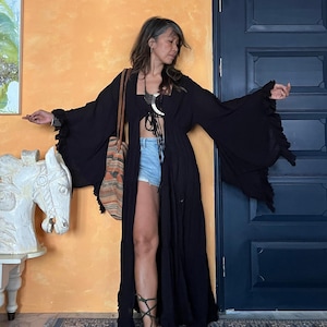 May include: A black, flowing kimono-style robe with wide sleeves. The robe is open in the front, revealing a black top and denim shorts. A patterned bag hangs from the shoulder. The background features an orange wall and a dark blue door.