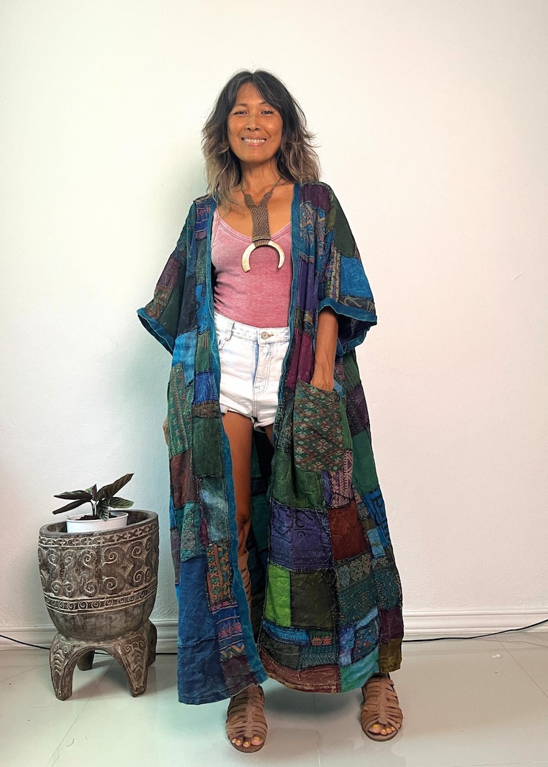 May include: A long, open-front patchwork duster coat featuring a variety of blue, green, and purple fabric squares with diverse patterns. The garment includes two large front pockets, three-quarter length sleeves, and a loose, flowing silhouette.