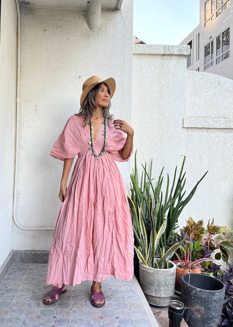 May include: A woman wearing a pink maxi dress with a wide neckline and a gathered skirt. She is wearing a straw hat and a green beaded necklace. She is standing on a tiled patio with plants in pots.