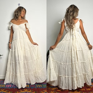 May include: Off-white, tiered maxi dress with an off-the-shoulder neckline and ruffled sleeves. The dress features a flowing, layered skirt and a tie-back detail. The fabric appears to be lightweight and textured, suitable for warm weather.
