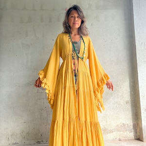 May include: A long, flowing yellow dress with ruffled sleeves and hem. The dress is open in the front and tied at the waist. The model is wearing a green top and a beaded necklace. The background is a light gray wall.