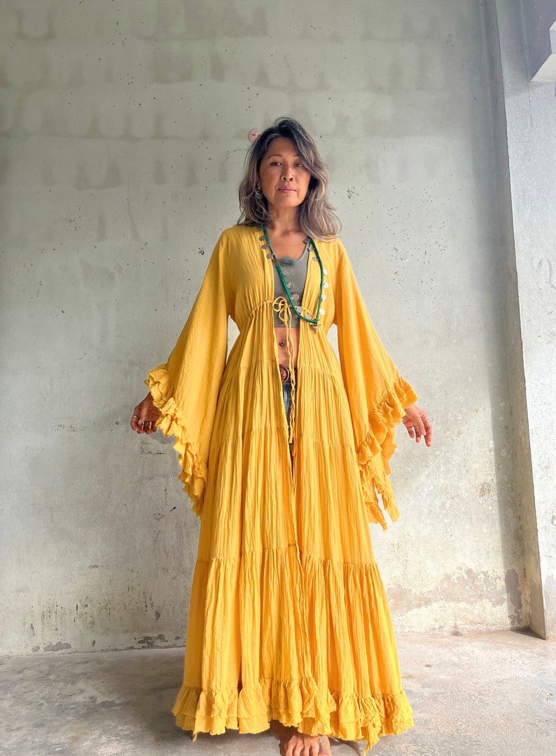 May include: A long, flowing yellow dress with ruffled sleeves and hem. The dress is open in the front and tied at the waist. The model is wearing a green top and a beaded necklace. The background is a light gray wall.