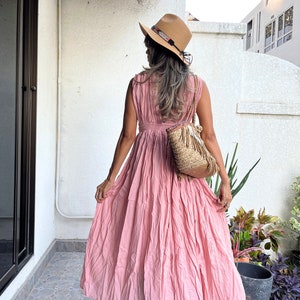 May include: A pink sleeveless maxi dress with a gathered skirt and a cinched waist. The dress is worn by a person wearing a straw hat with a brown band and a woven bag.