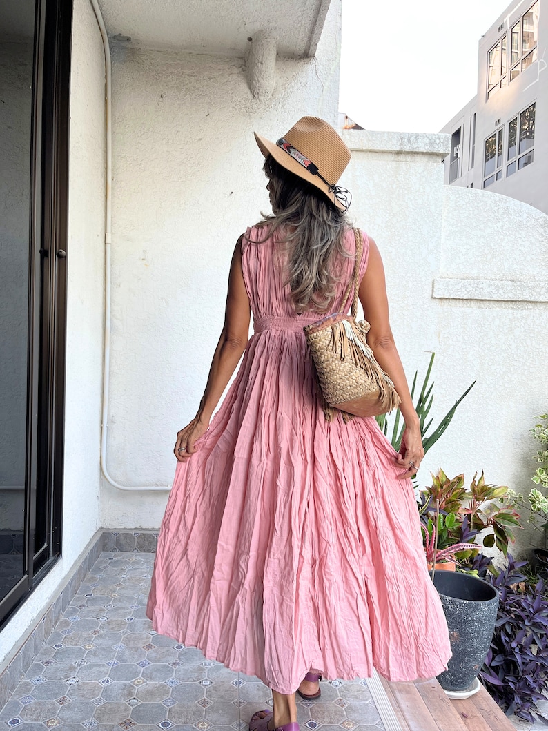 May include: A pink sleeveless maxi dress with a gathered skirt and a cinched waist. The dress is worn by a person wearing a straw hat with a brown band and a woven bag.