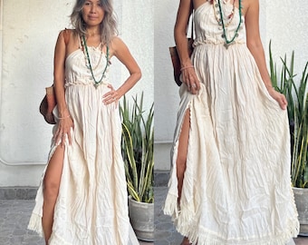 May include: A white maxi dress with a fringe hem and a halter neckline. The dress is made of a lightweight fabric and has a relaxed fit. The model is wearing the dress with a layered necklace and sandals.