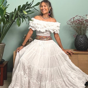 May include: A white off-the-shoulder peasant top with a ruffled neckline and a white tiered maxi skirt. The skirt has a colorful embroidered belt with silver accents.