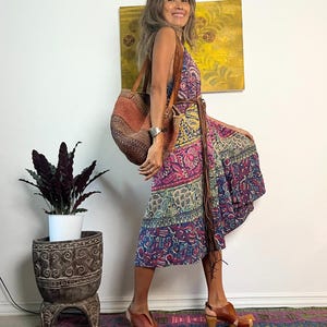 May include: A woman wearing a colorful patterned wrap dress with a brown leather belt and a brown woven tote bag. She is standing on a patterned rug with a potted plant in the background.