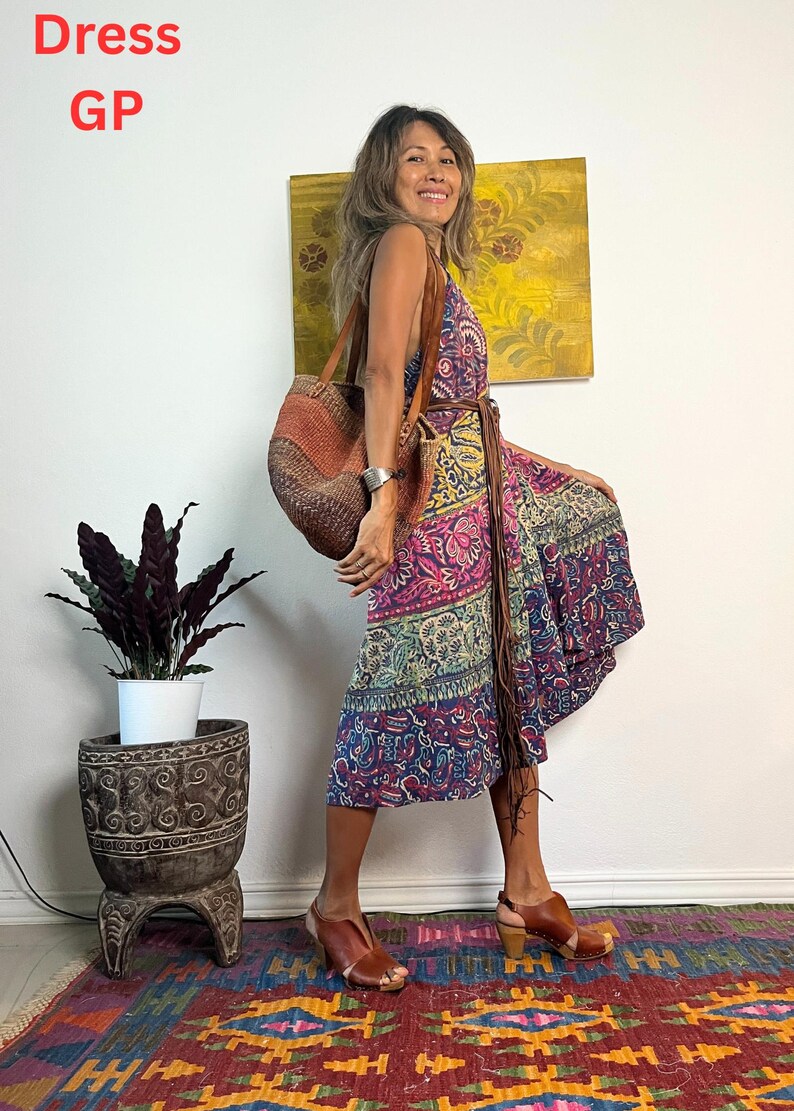 May include: A woman wearing a colorful patterned wrap dress with a brown leather belt and a brown woven tote bag. She is standing on a patterned rug with a potted plant in the background.