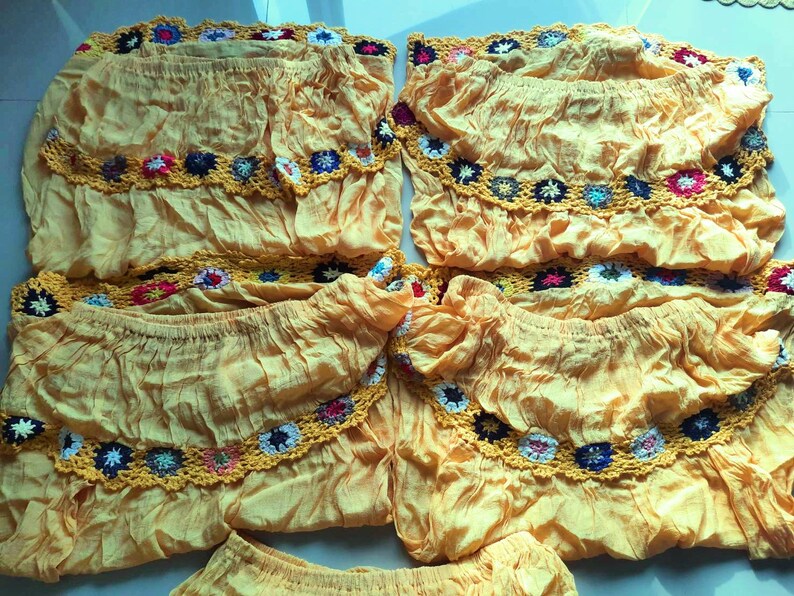 May include: Five yellow off-the-shoulder tops with crocheted trim in various colors. The tops are made of a lightweight fabric and have a ruffled design.
